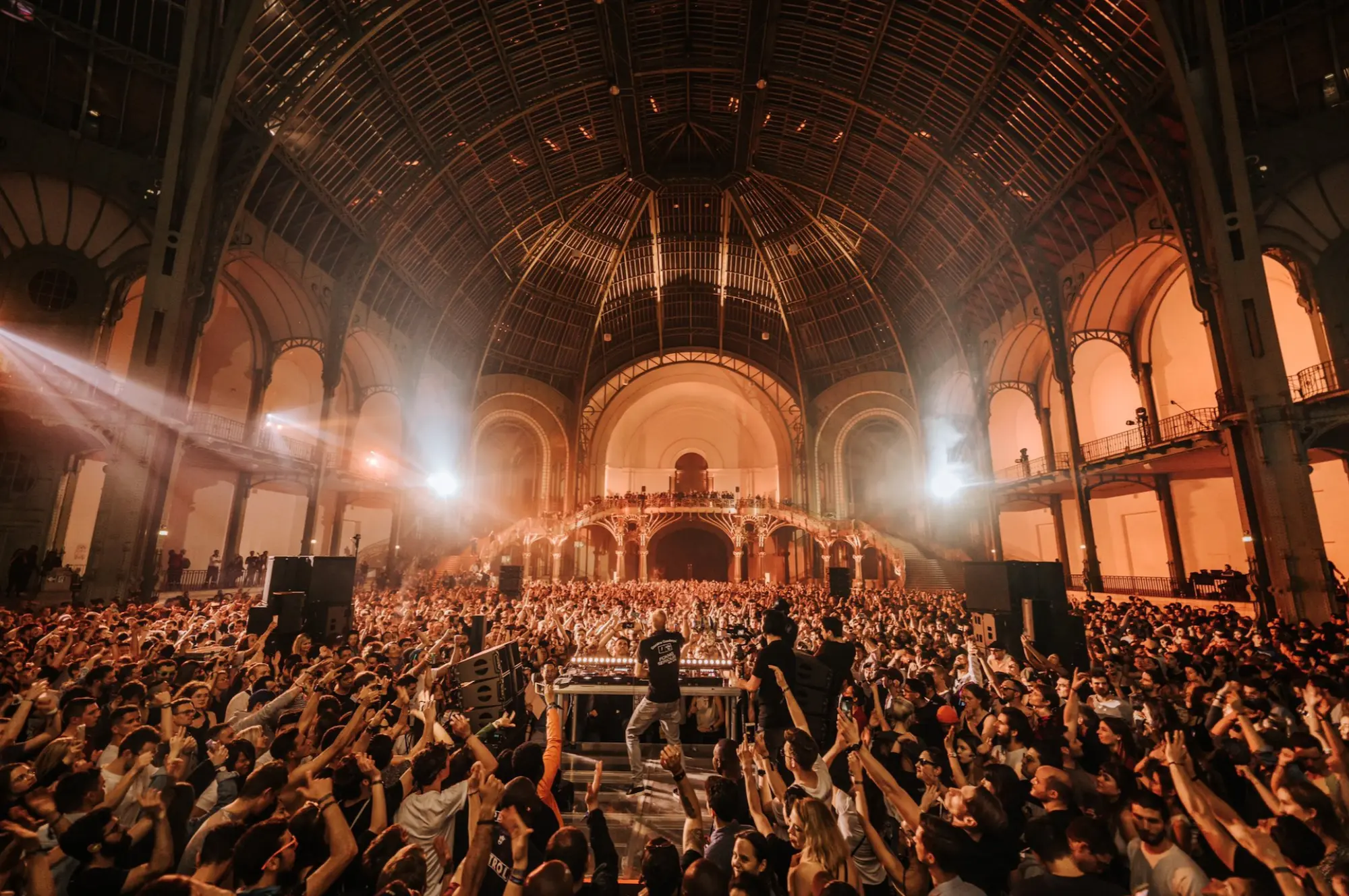 A packed indoor crowd around a central DJ booth beneath a vast iron-and-glass domed heritage hall, lit by dramatic beams of light.