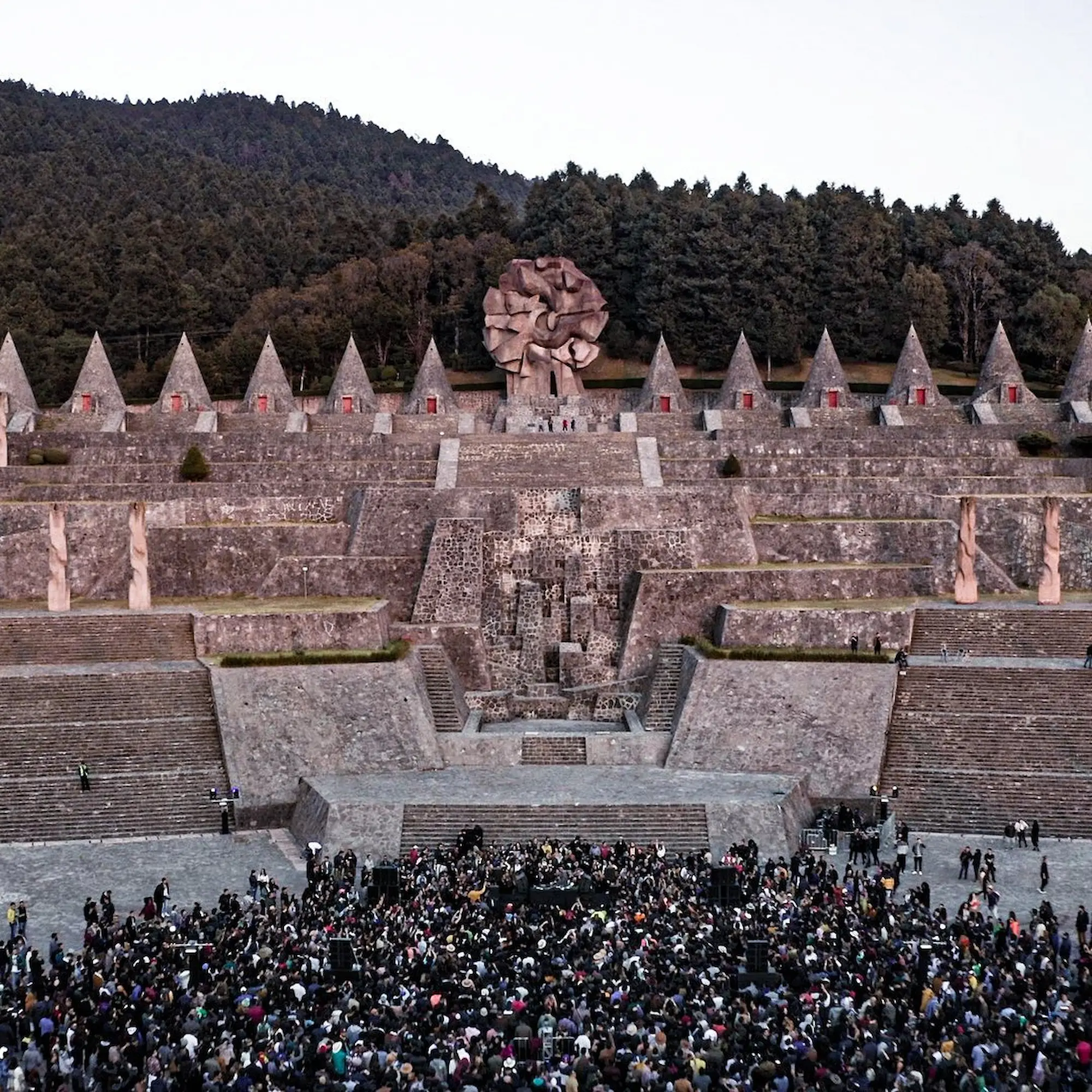 A crowd gathered before a monumental terraced stone amphitheatre at dusk, framed by forested hills.