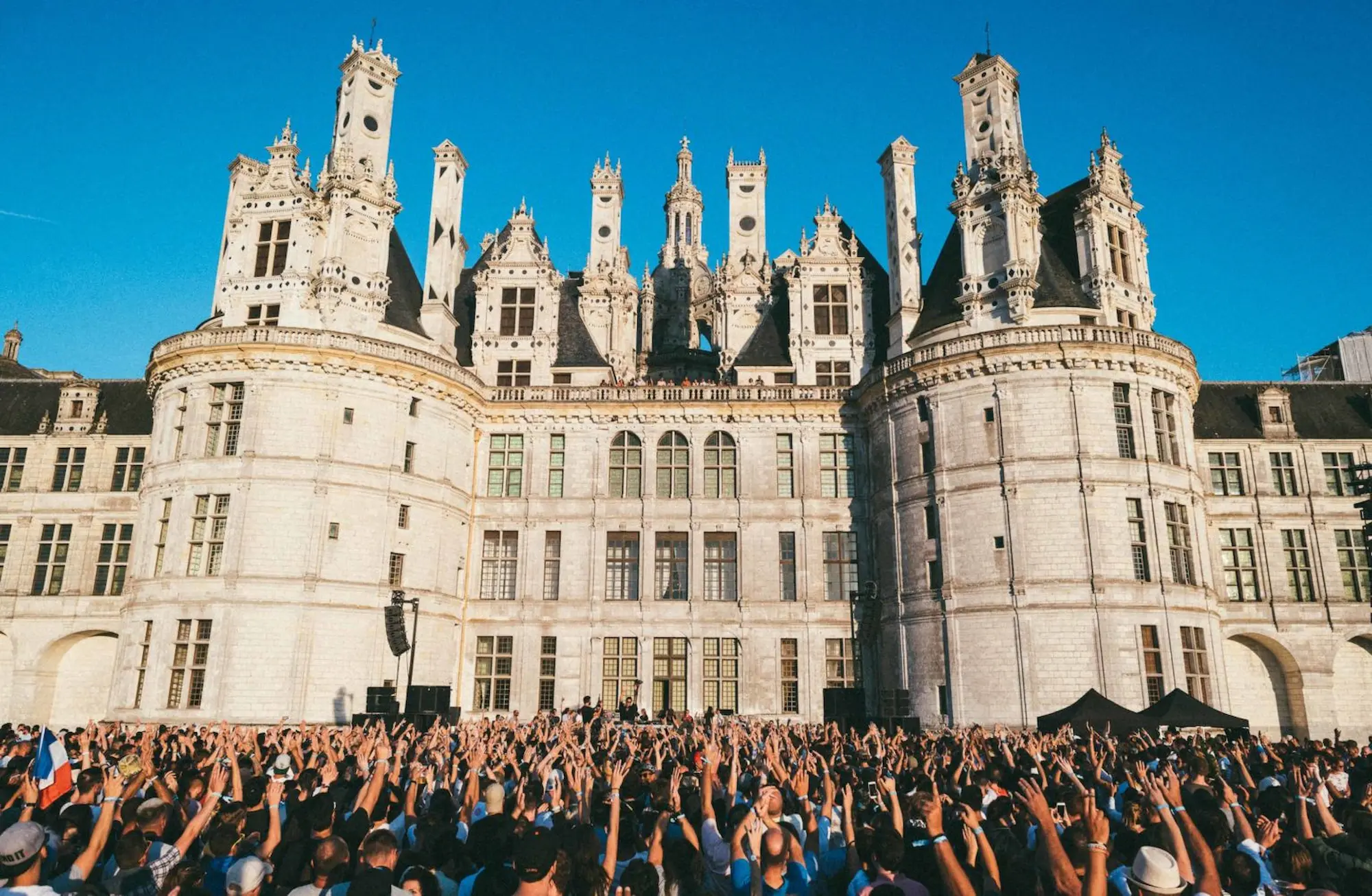 A large crowd with raised hands in front of the Renaissance towers of the Château de Chambord under a clear blue sky.