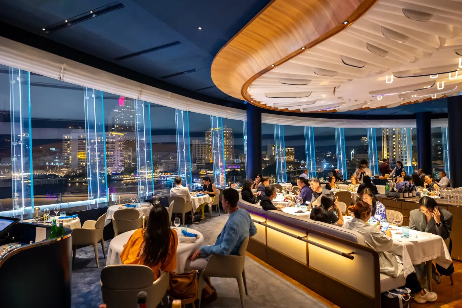A full dining room at Blue by Alain Ducasse at night, with the lights of the far bank of the Chao Phraya in the windows