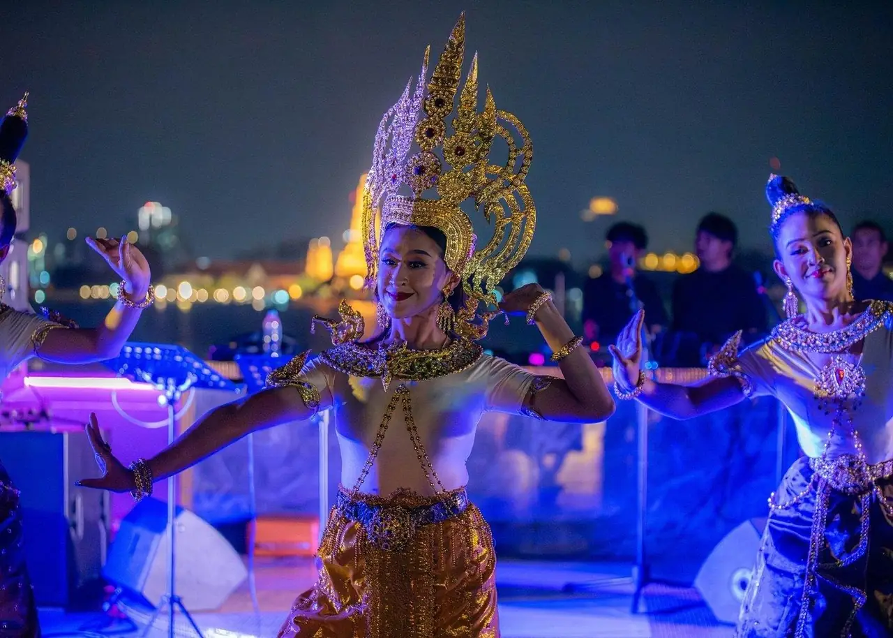 Thai classical dancers in golden headdresses on deck, the lit riverbank behind them