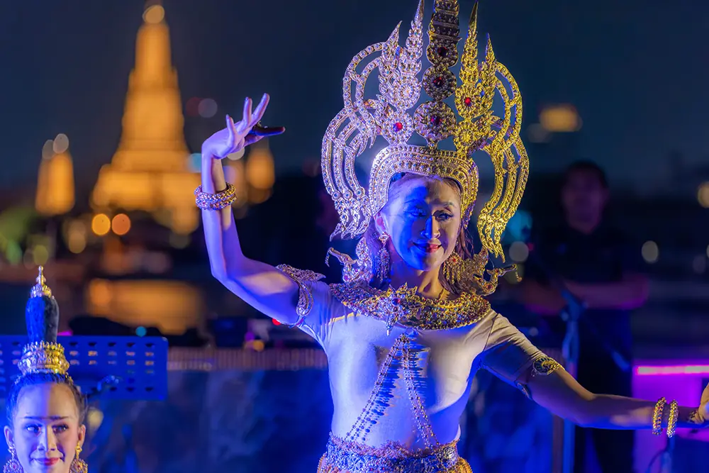 A Thai classical dancer under blue light on deck, Wat Arun glowing gold behind her