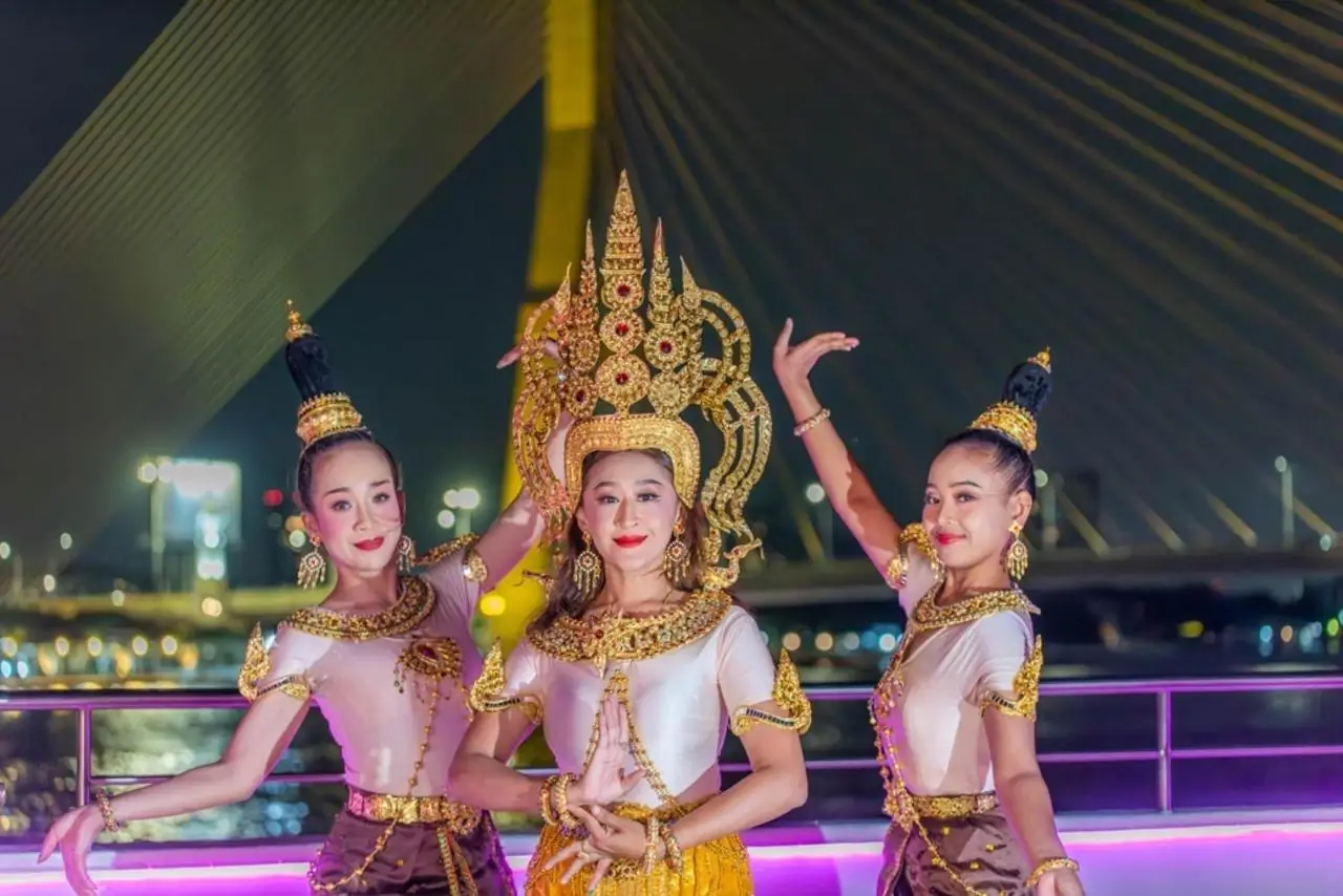 Three Thai dancers in gold headdresses on deck, a lit bridge behind them
