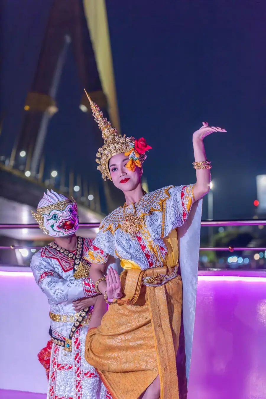A Thai dancer and a masked Khon performer posing on deck under a lit bridge