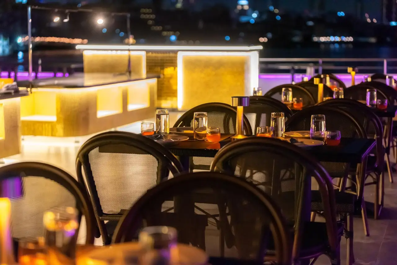 Tables set on the rooftop deck beside a softly lit counter, the river behind