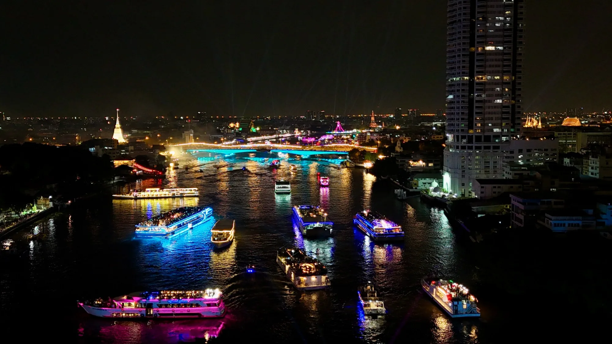 A fleet of lit-up dinner cruise boats sailing on the Chao Phraya River in Bangkok at night