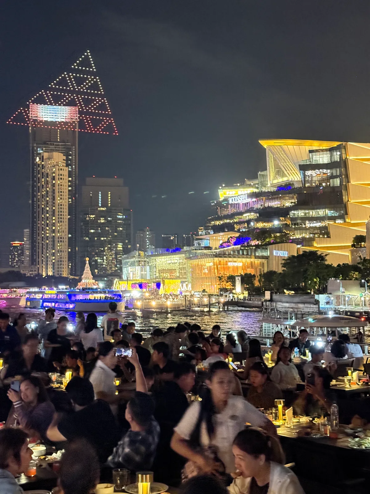 Dinner tables by the Chao Phraya at night, looking across the river to ICONSIAM during a drone light show