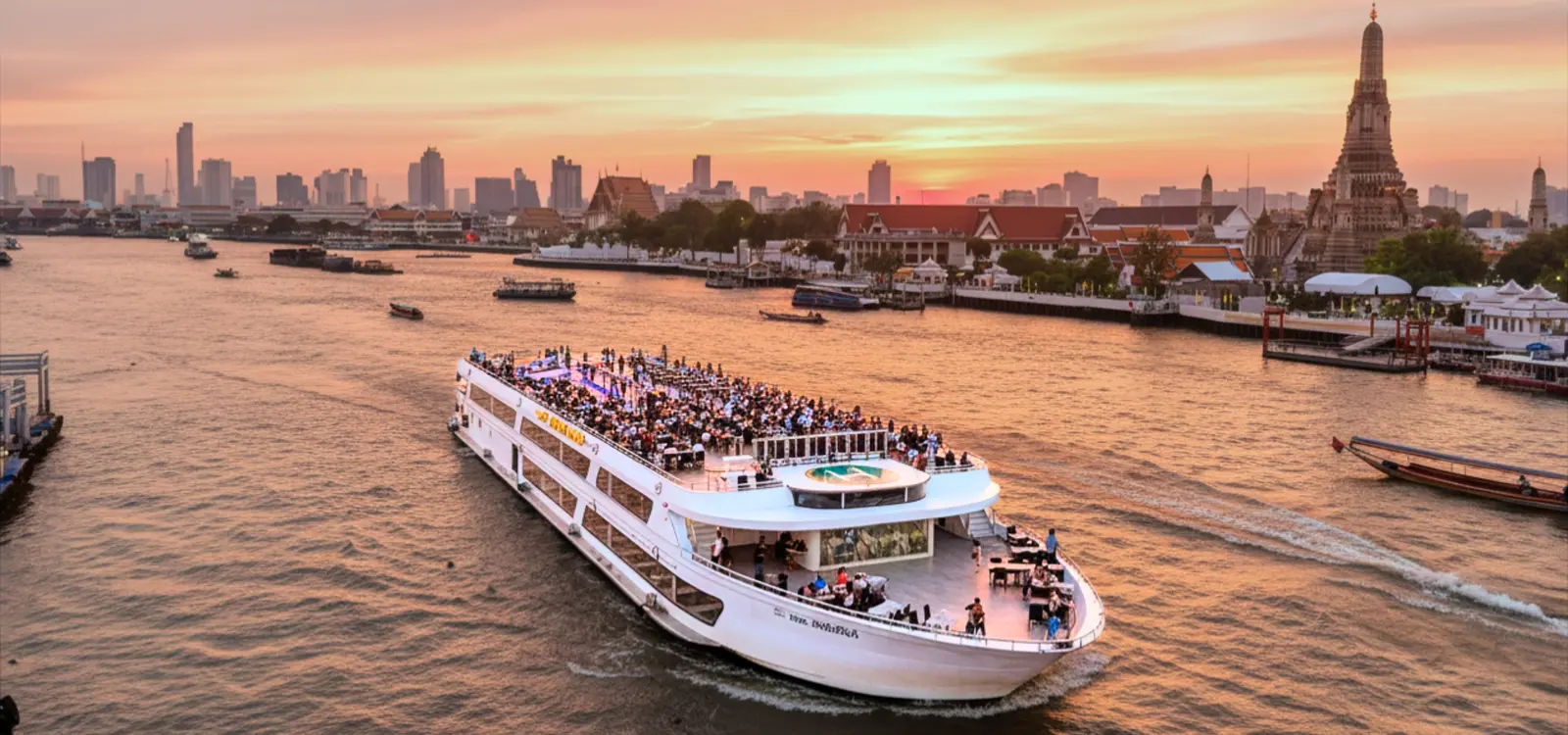 The Luxury White sailing past Wat Arun at sunset, its rooftop deck full of diners