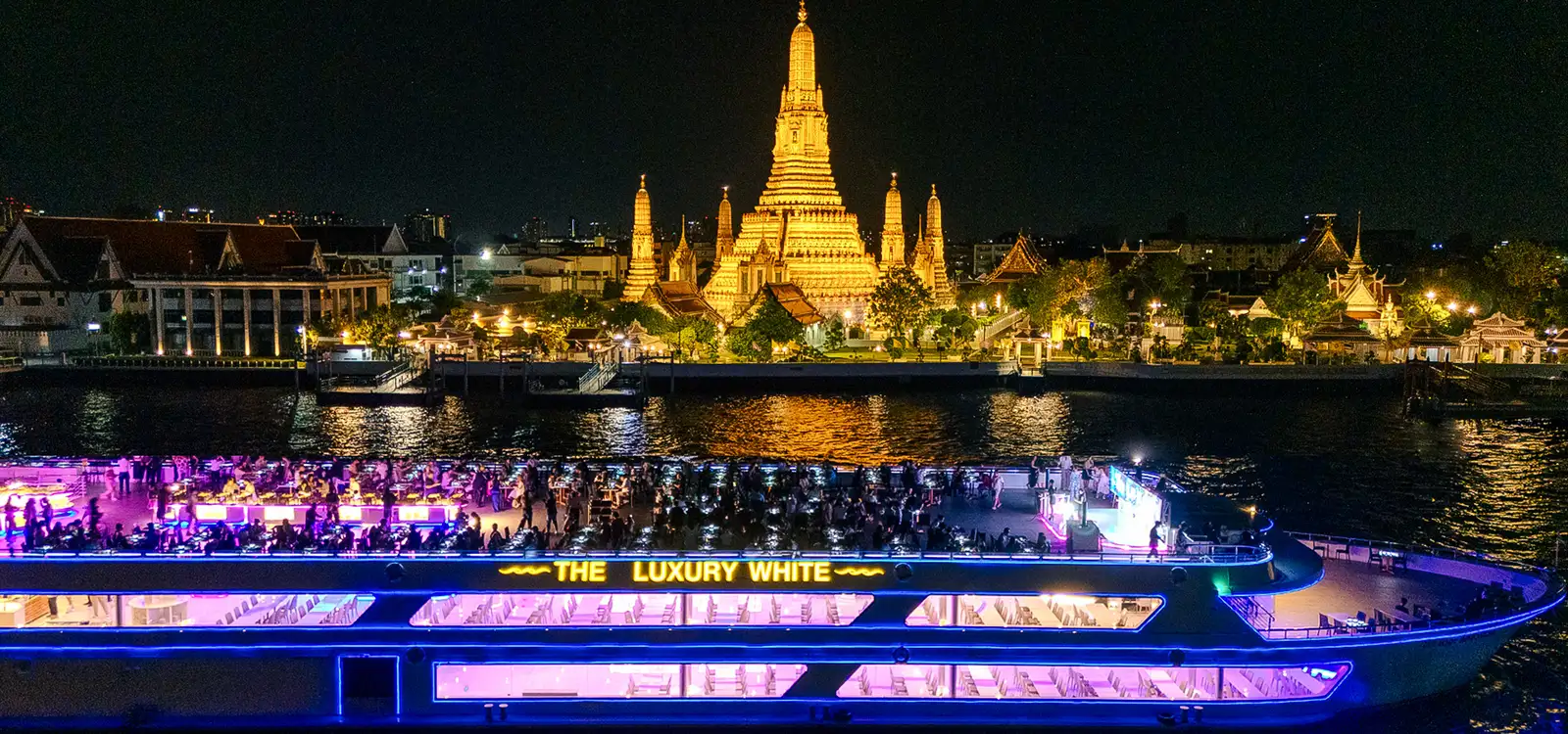 The Luxury White lit up in blue and pink on the river at night, with Wat Arun behind