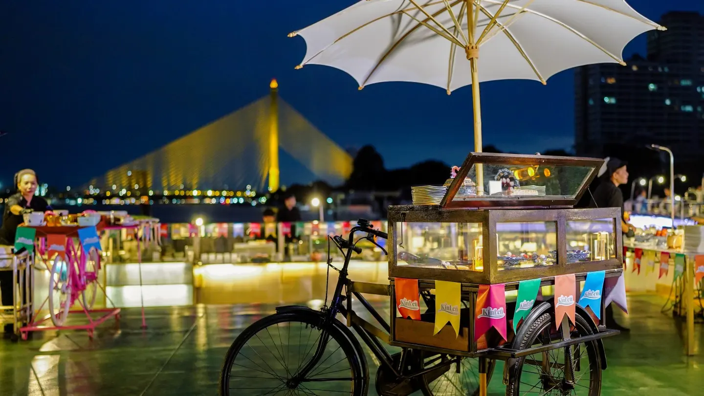 A street-food cart on the open deck of a Riverside cruise boat, with the Rama VIII Bridge behind