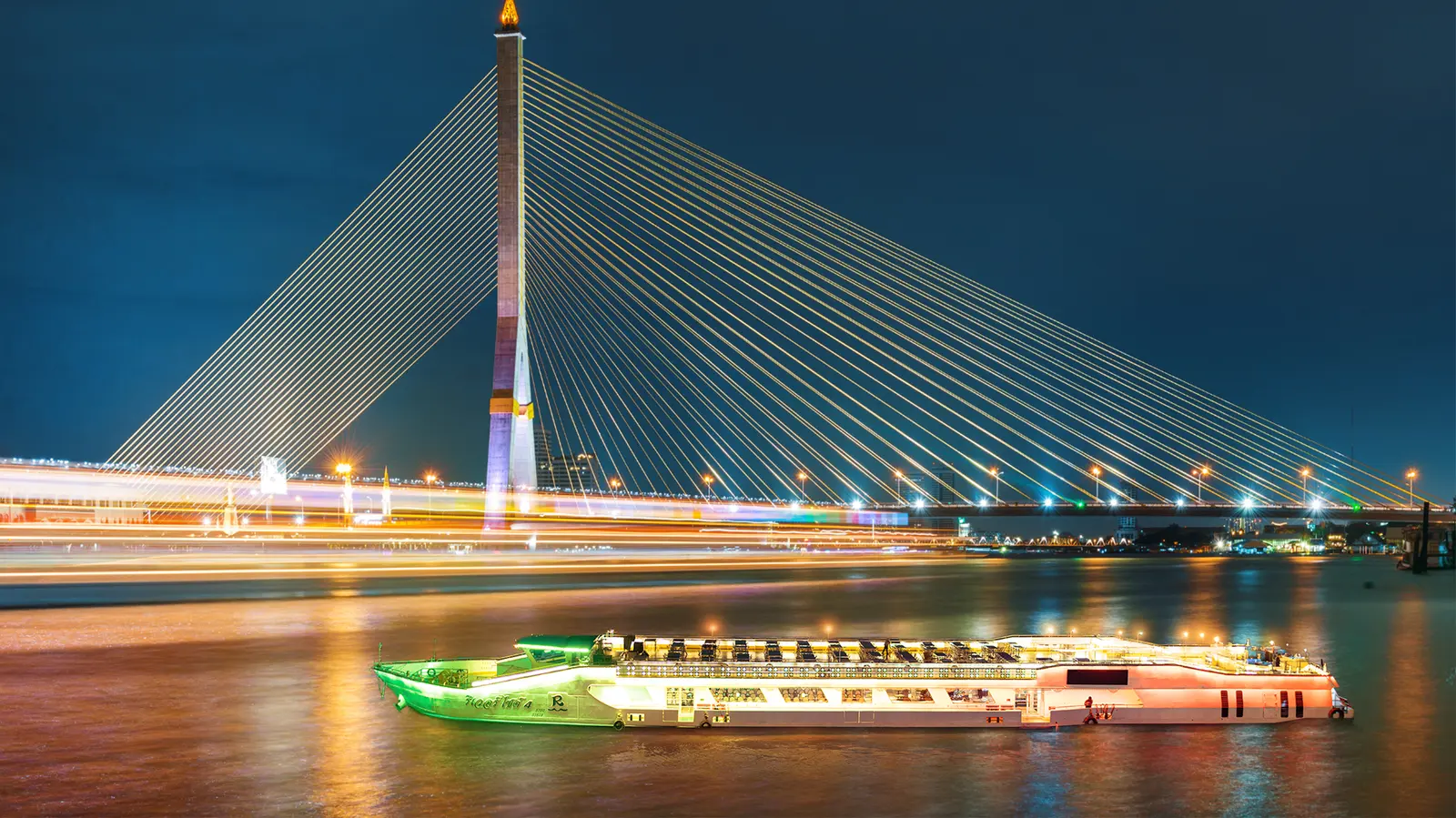 A Riverside Bangkok cruise boat on the river at night, in front of the Rama VIII Bridge
