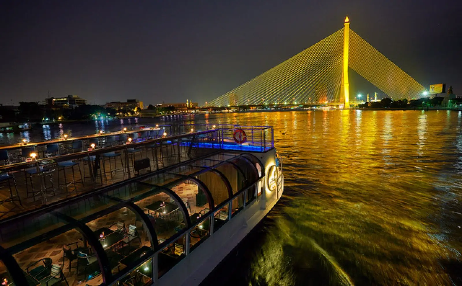 The lit rooftop deck of the Saffron Cruise at night, with the golden Rama VIII Bridge ahead