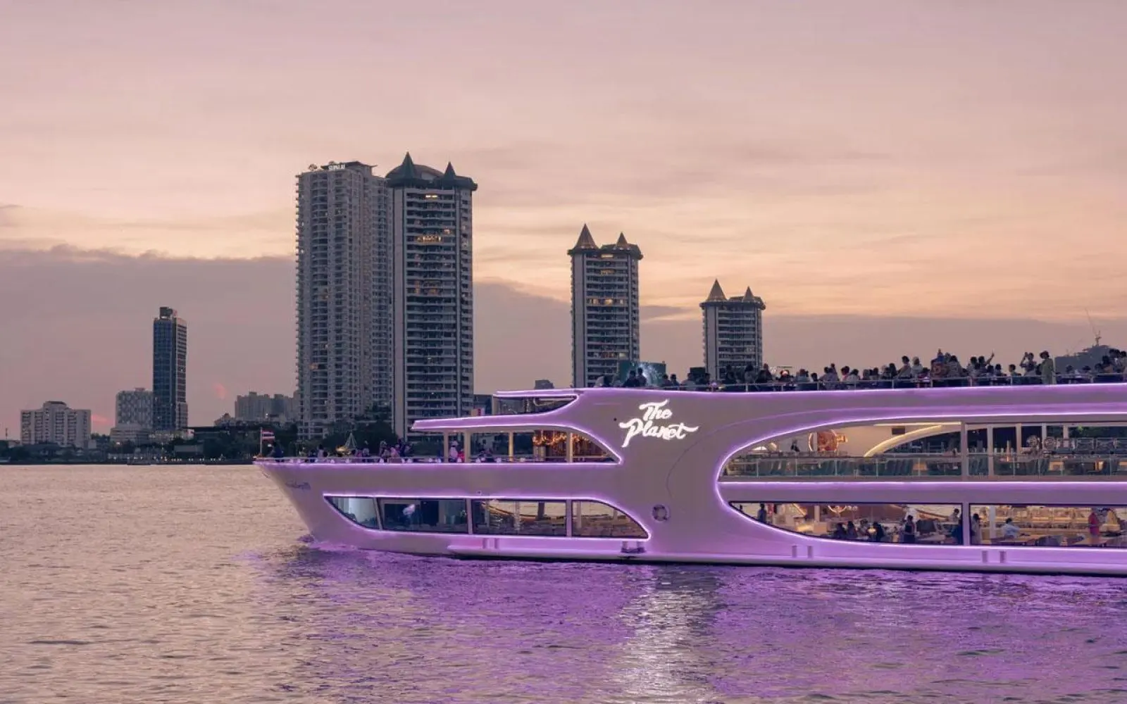 The Planet Cruise at dusk, with guests on its rooftop deck and riverside towers behind
