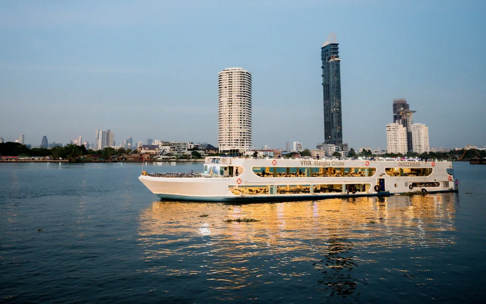 The VIVA Alangka Cruise boat lit up on the Chao Phraya at dusk, riverside towers behind