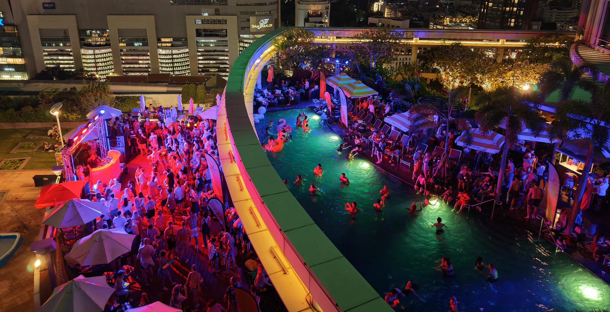 Rooftop pool party at night in Bangkok — a crowd around a lit pool with neon lights and the city skyline behind