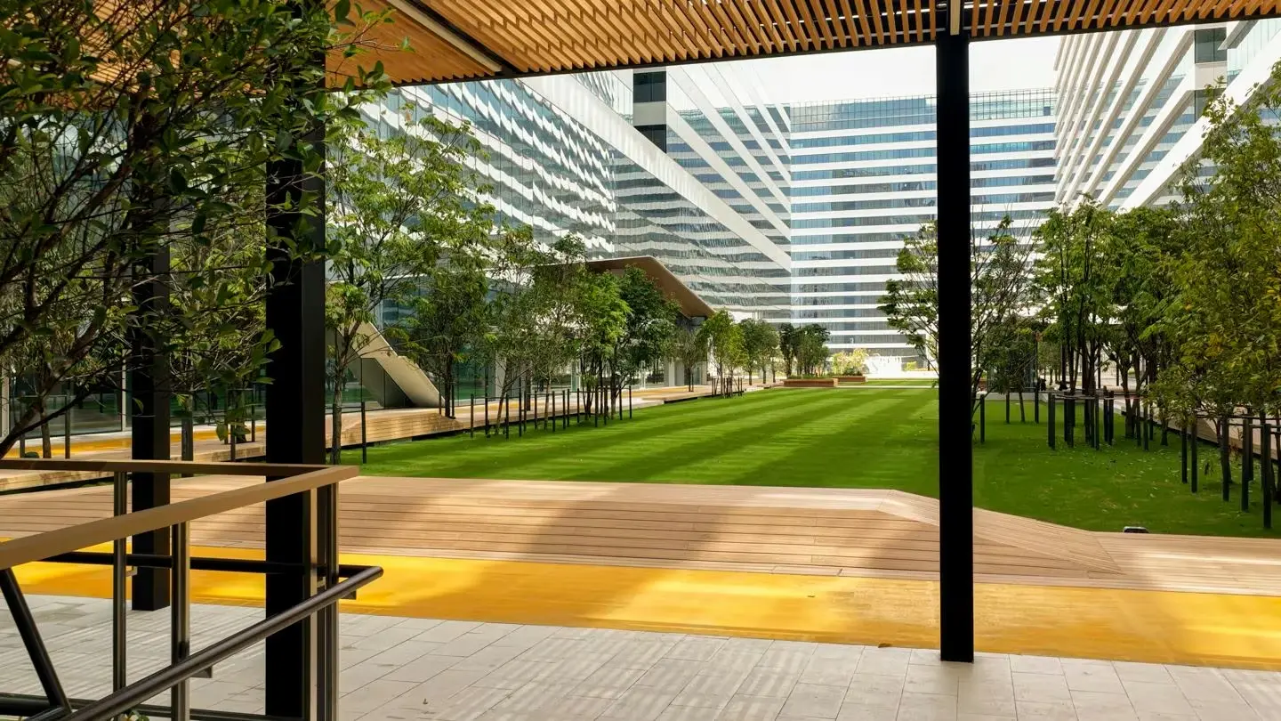 Cloud 11 Park by day: a long green lawn and young trees between glass office towers, seen from under a wooden pergola