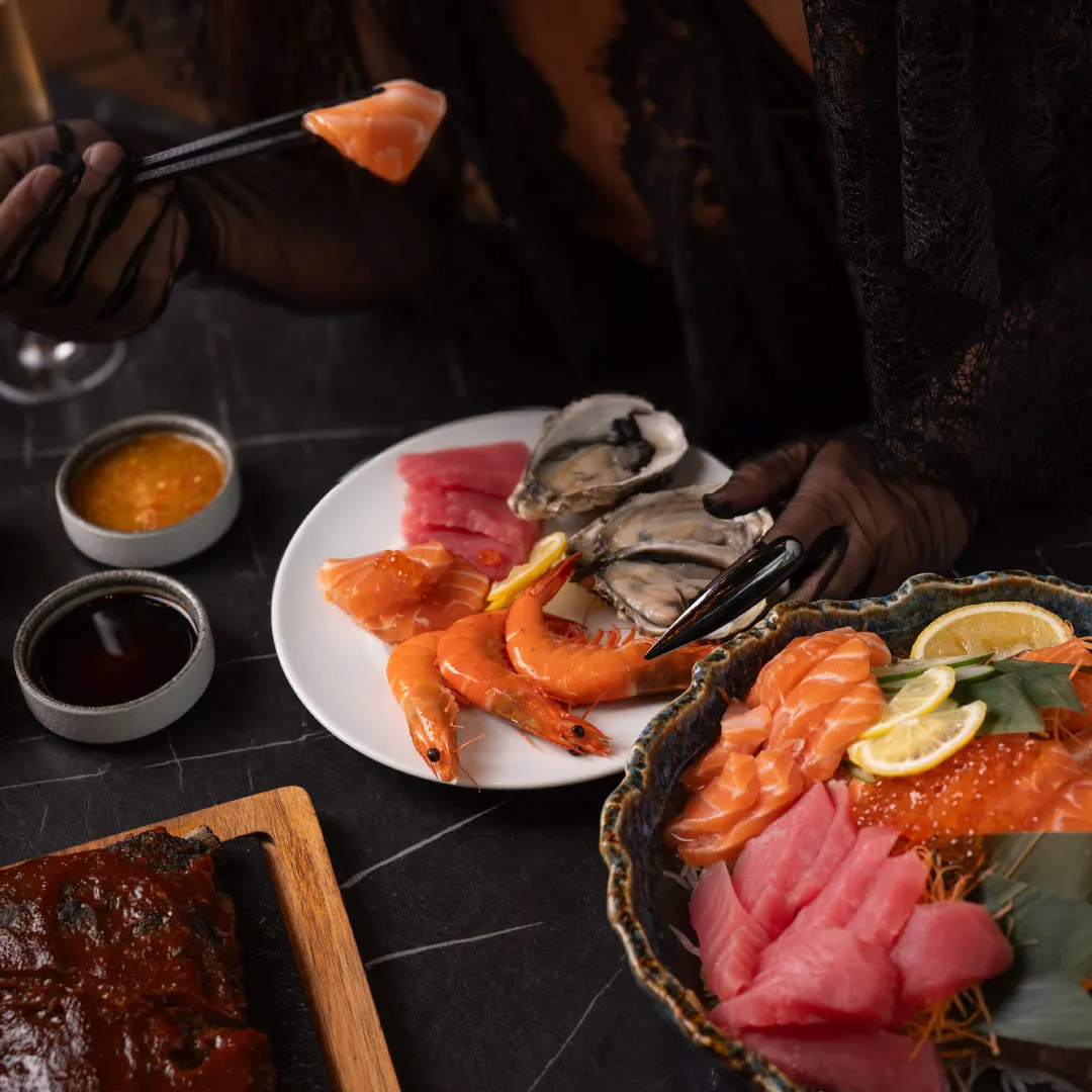 Salmon and tuna sashimi, prawns and oysters on a dark marble table at W Does Brunch, a hand in black lace gloves holding chopsticks