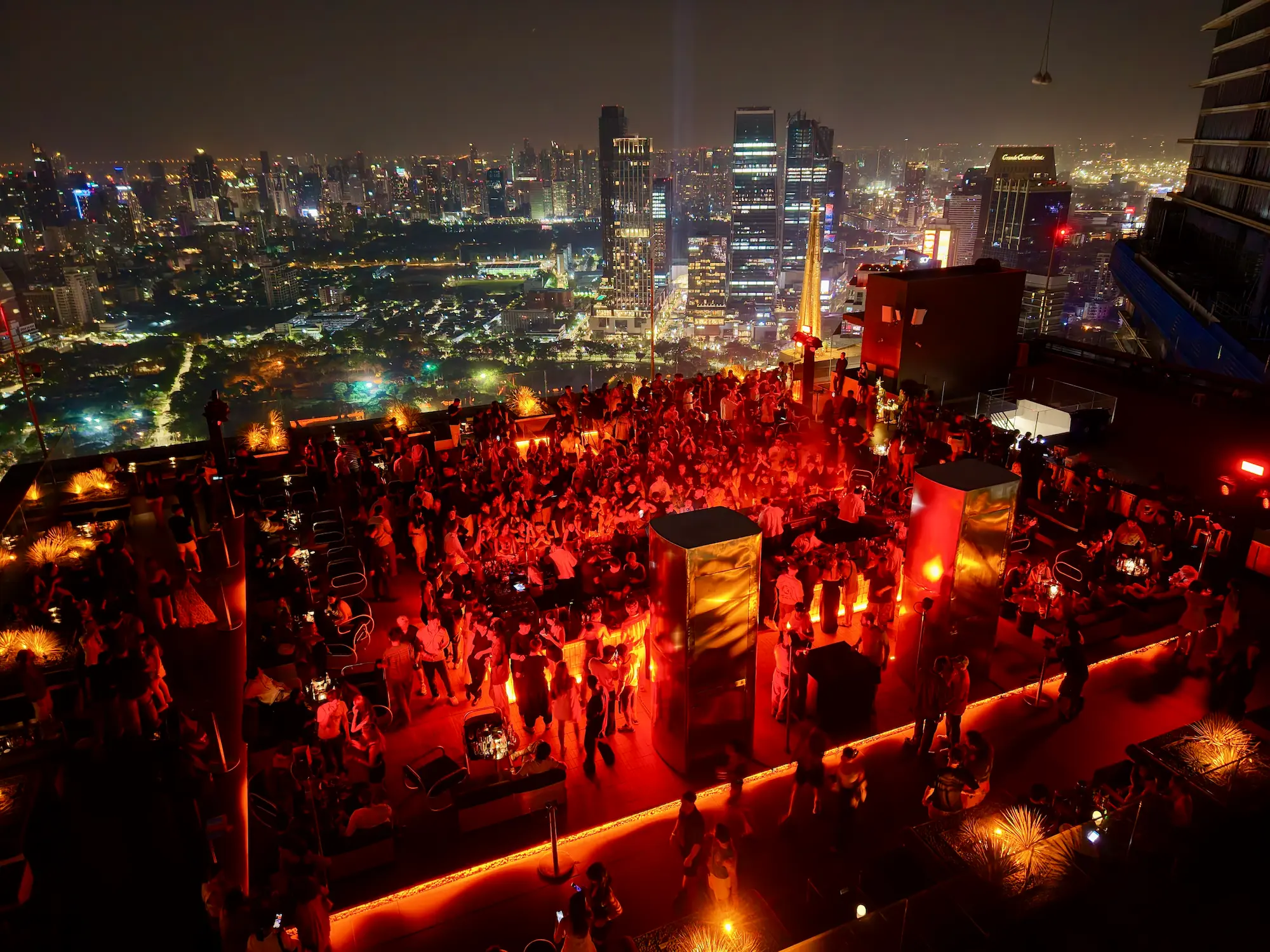 A large crowd partying on Aether's red-lit rooftop deck at night, the Bangkok skyline and Lumpini Park lit up behind