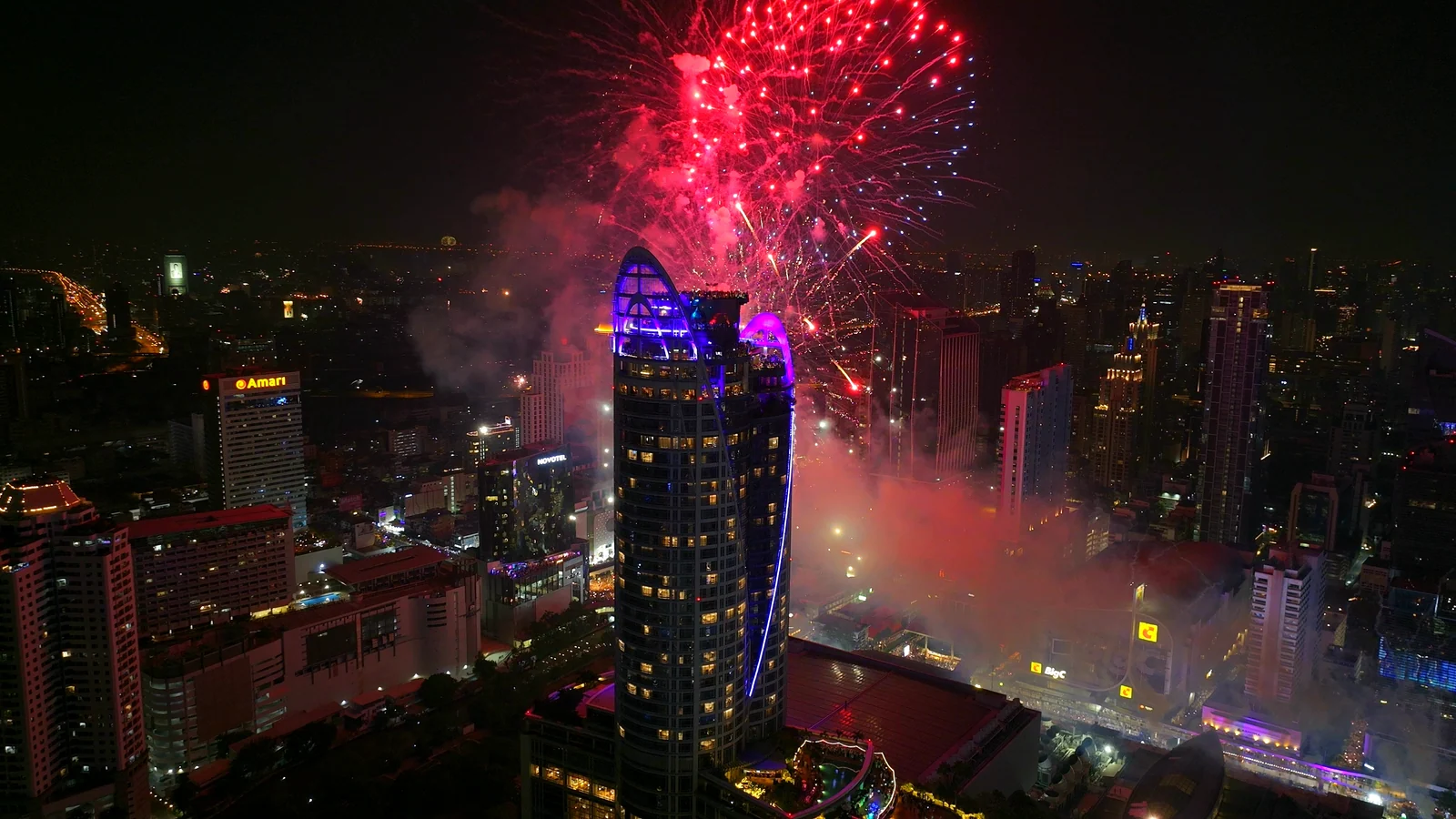 Pink fireworks bursting beside the blue-lit Centara Grand tower above the CentralWorld countdown at midnight, seen from the air