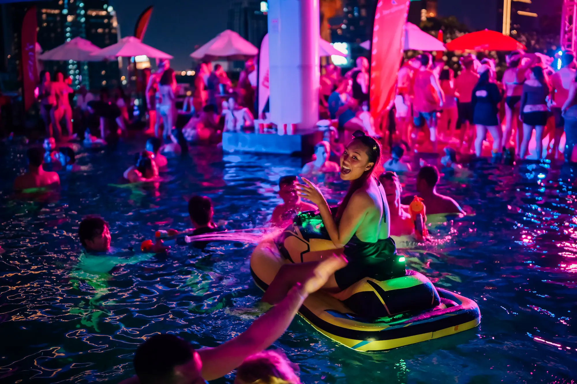 Night rooftop pool party in Bangkok, a crowd in a neon-lit pool with the city skyline behind