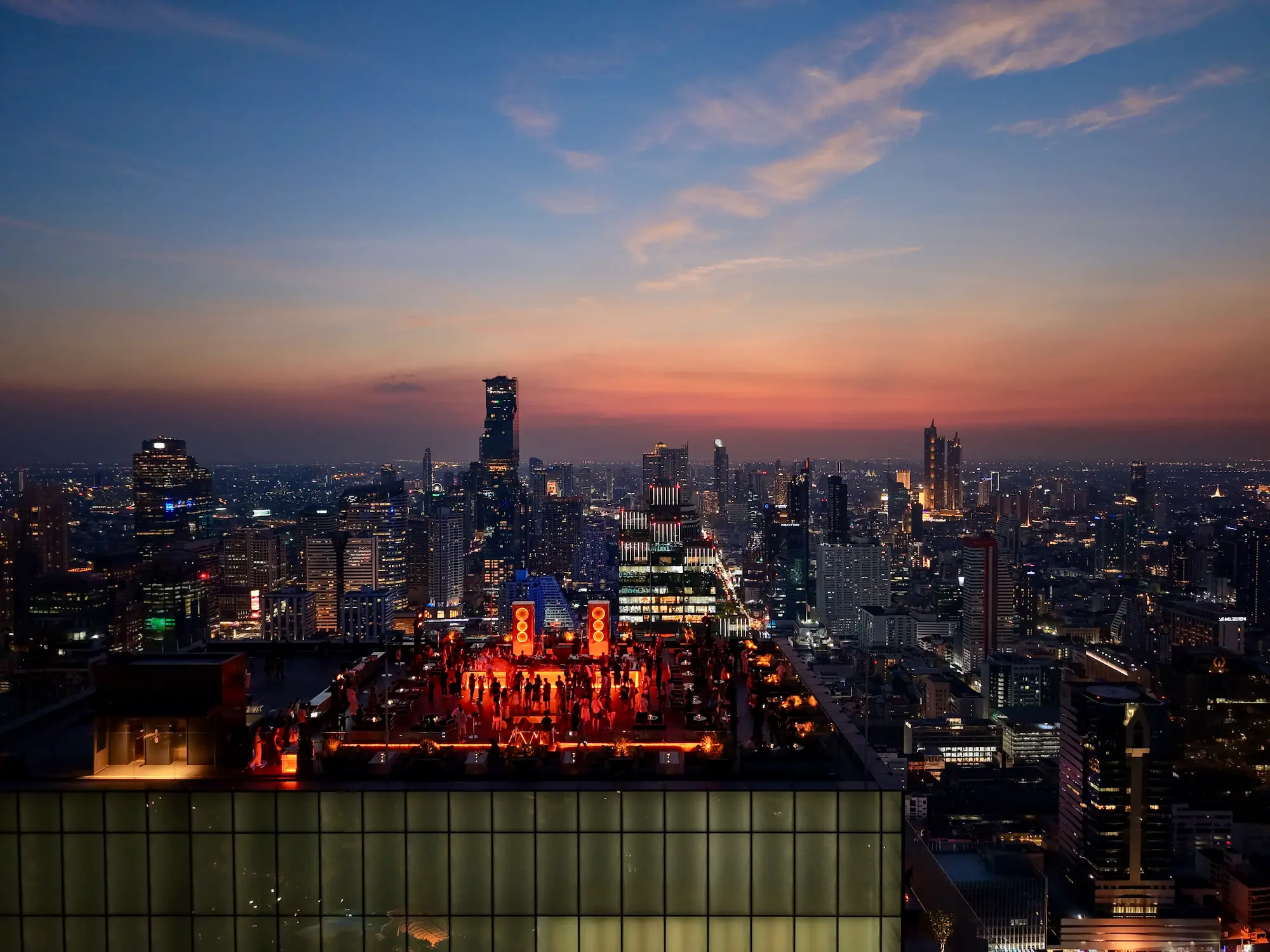 Aerial view of Aether's orange-lit rooftop bar high above Silom, the skyline behind it as dusk fades to blue