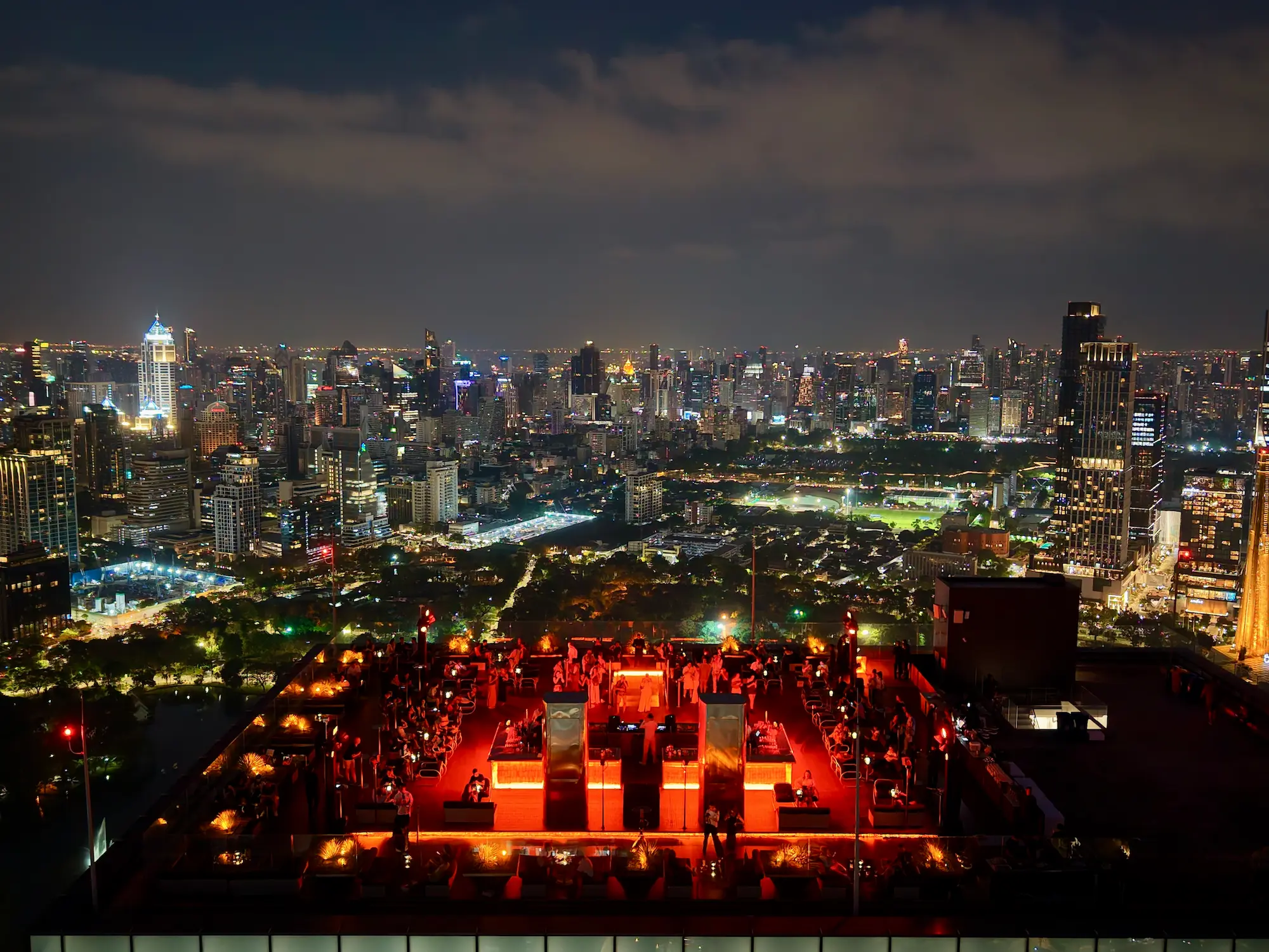 Aerial view of Aether rooftop bar at night, a red-lit DJ stage and crowd overlooking the Bangkok skyline and Lumpini Park