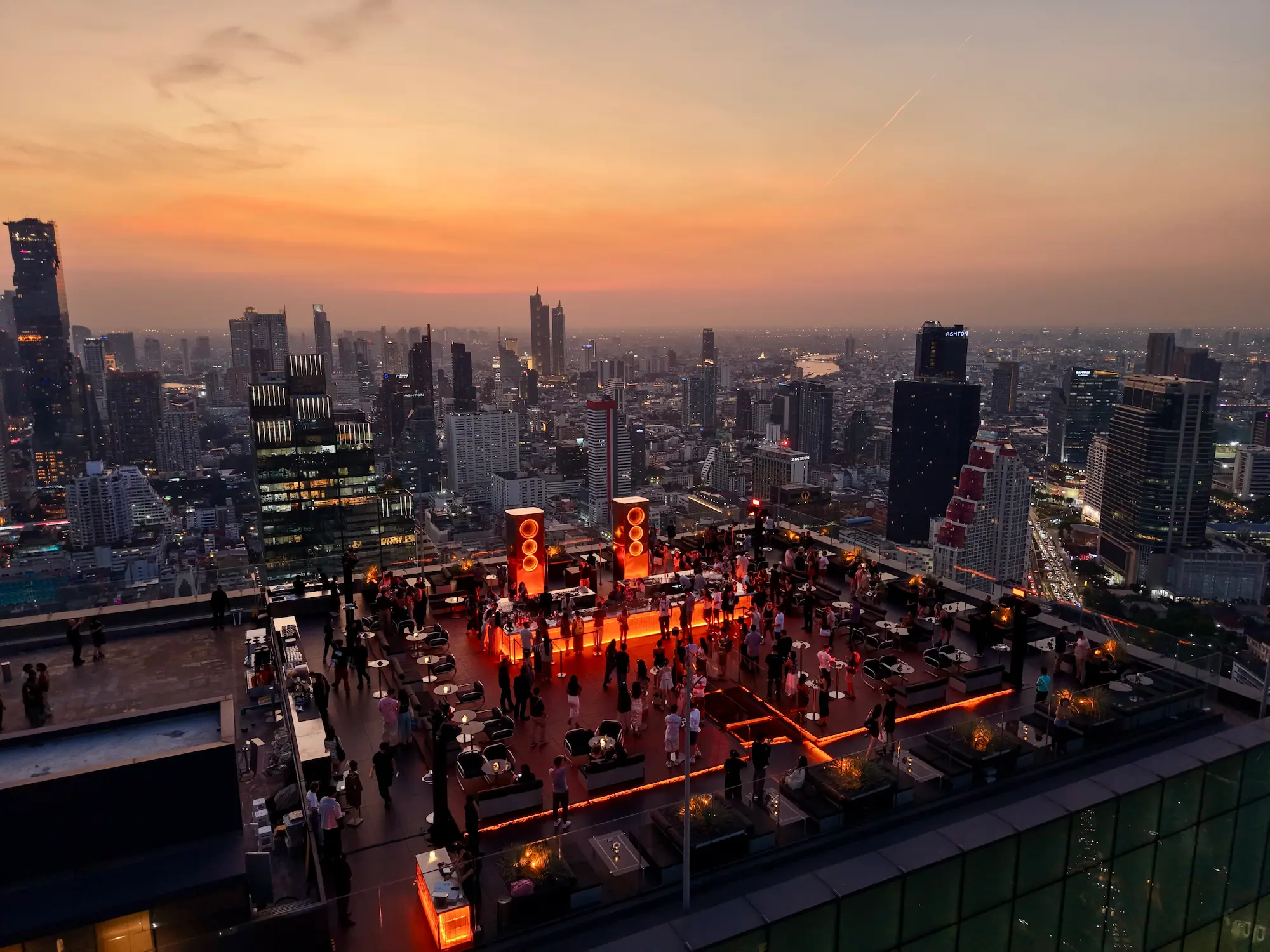 Aerial view of Aether rooftop bar at sunset, crowd and DJ under a vivid orange sky over the Bangkok skyline