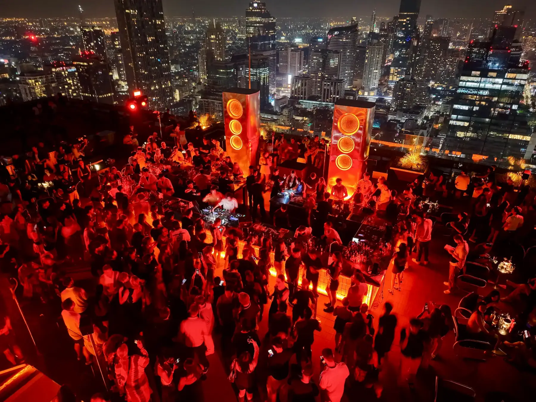 A packed crowd dancing around the DJ booth at Aether at night, glowing orange speaker towers and the Bangkok skyline behind