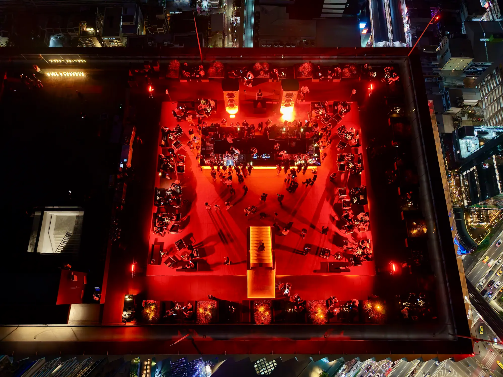 Top-down aerial of Aether's rooftop deck glowing red at night, the central bar and crowd surrounded by the dark city