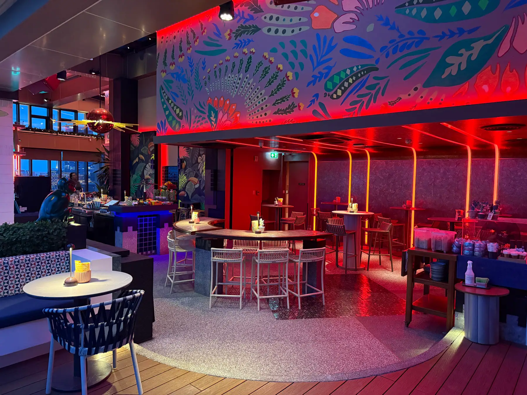 The lounge inside Bar.Yard under a large tropical mural, round tables and high stools in red light