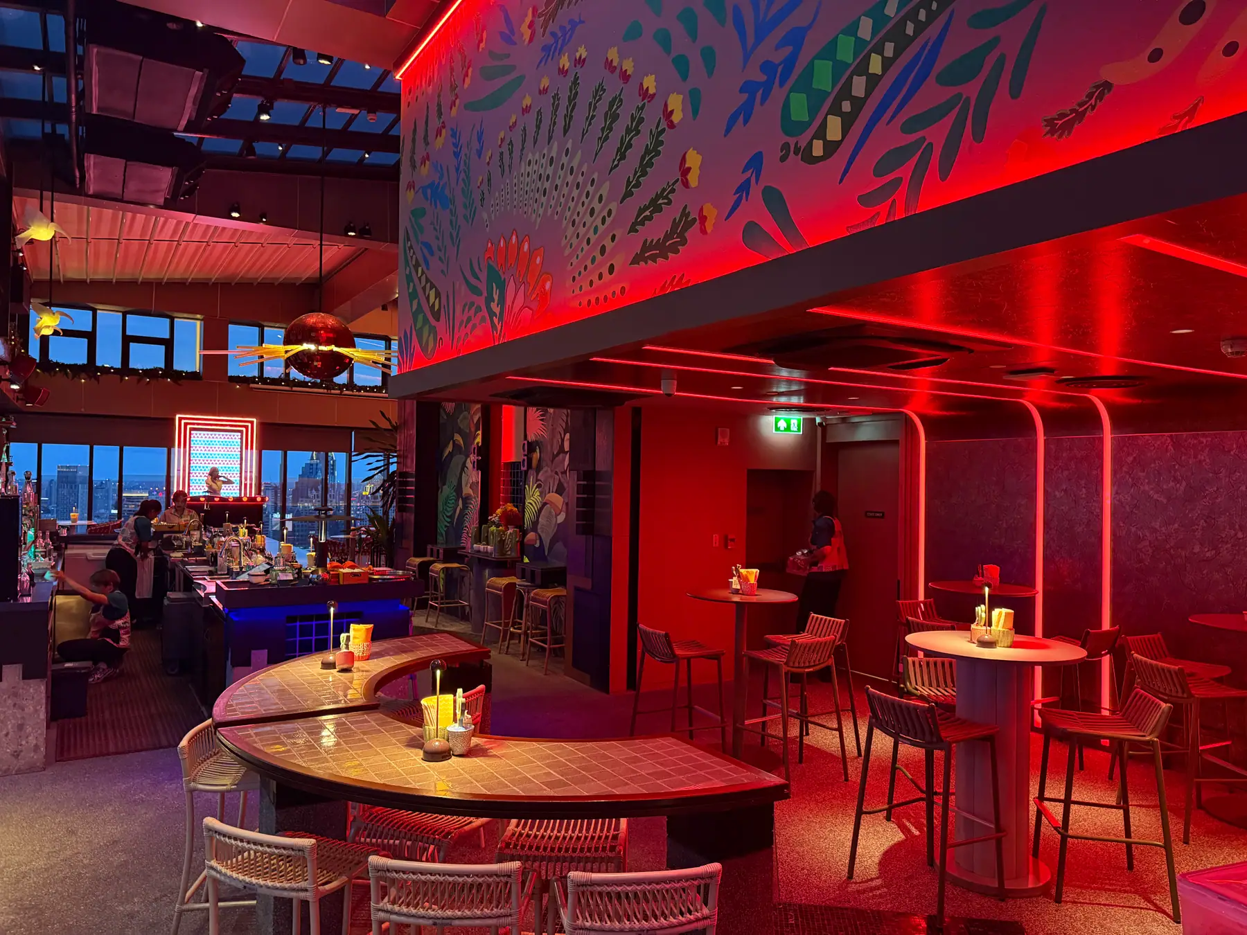 The red-lit interior of Bar.Yard: the bar on the left, a curved counter and stools in front, under the big tropical mural