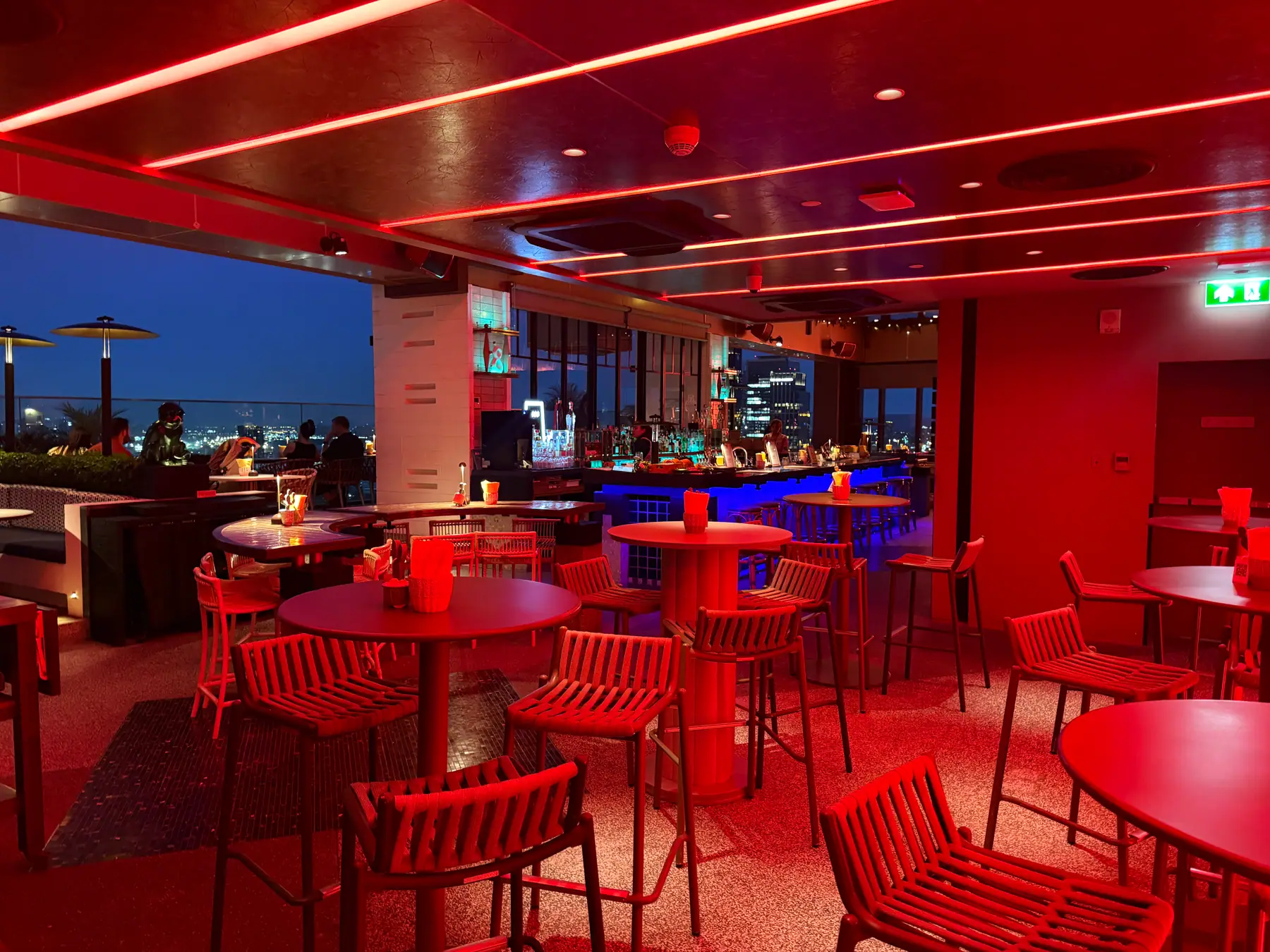 Red tables and chairs at Bar.Yard with the city lights through the windows behind