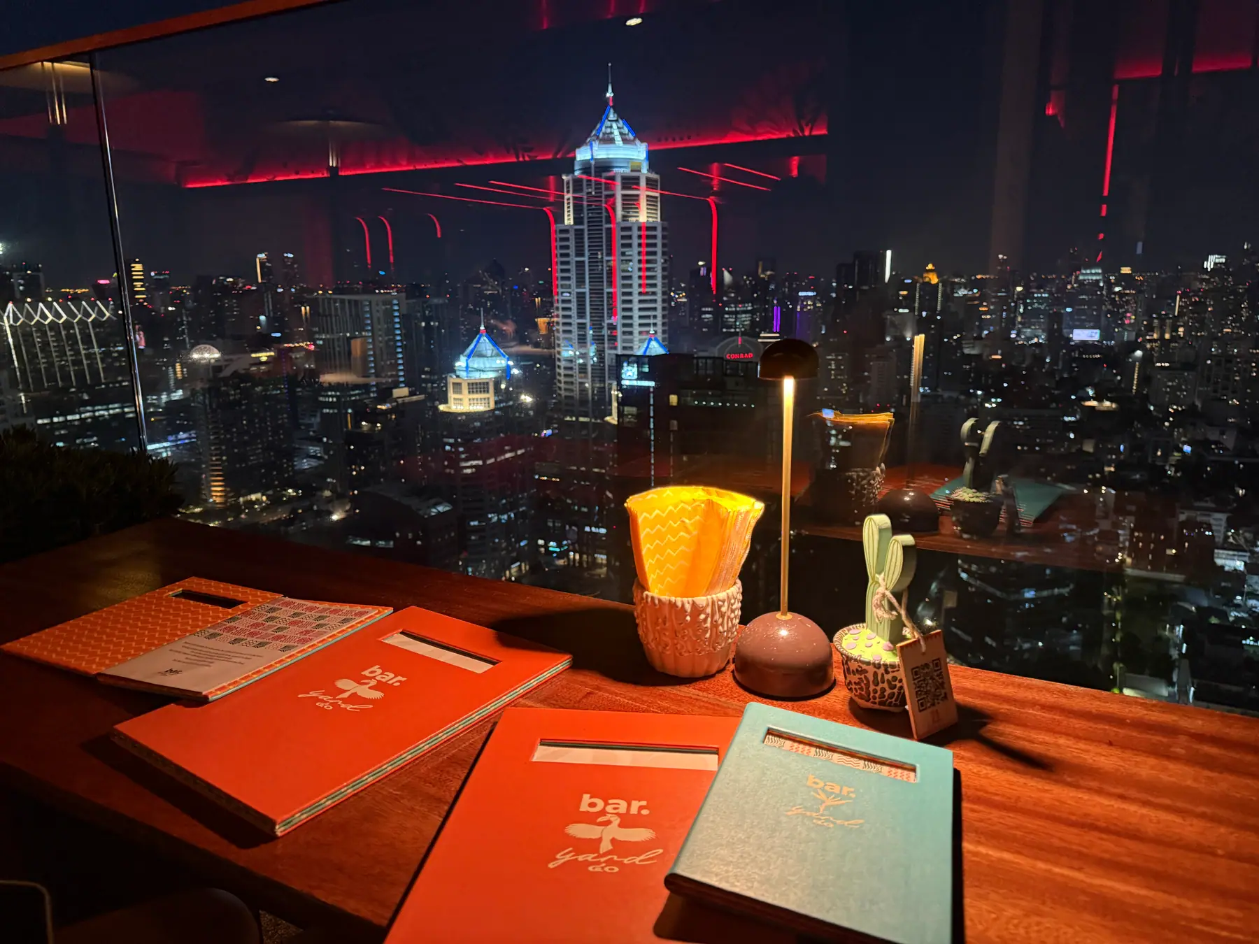 A window table at Bar.Yard at night, menus and a candle in front of the lit towers of the Bangkok skyline to the east