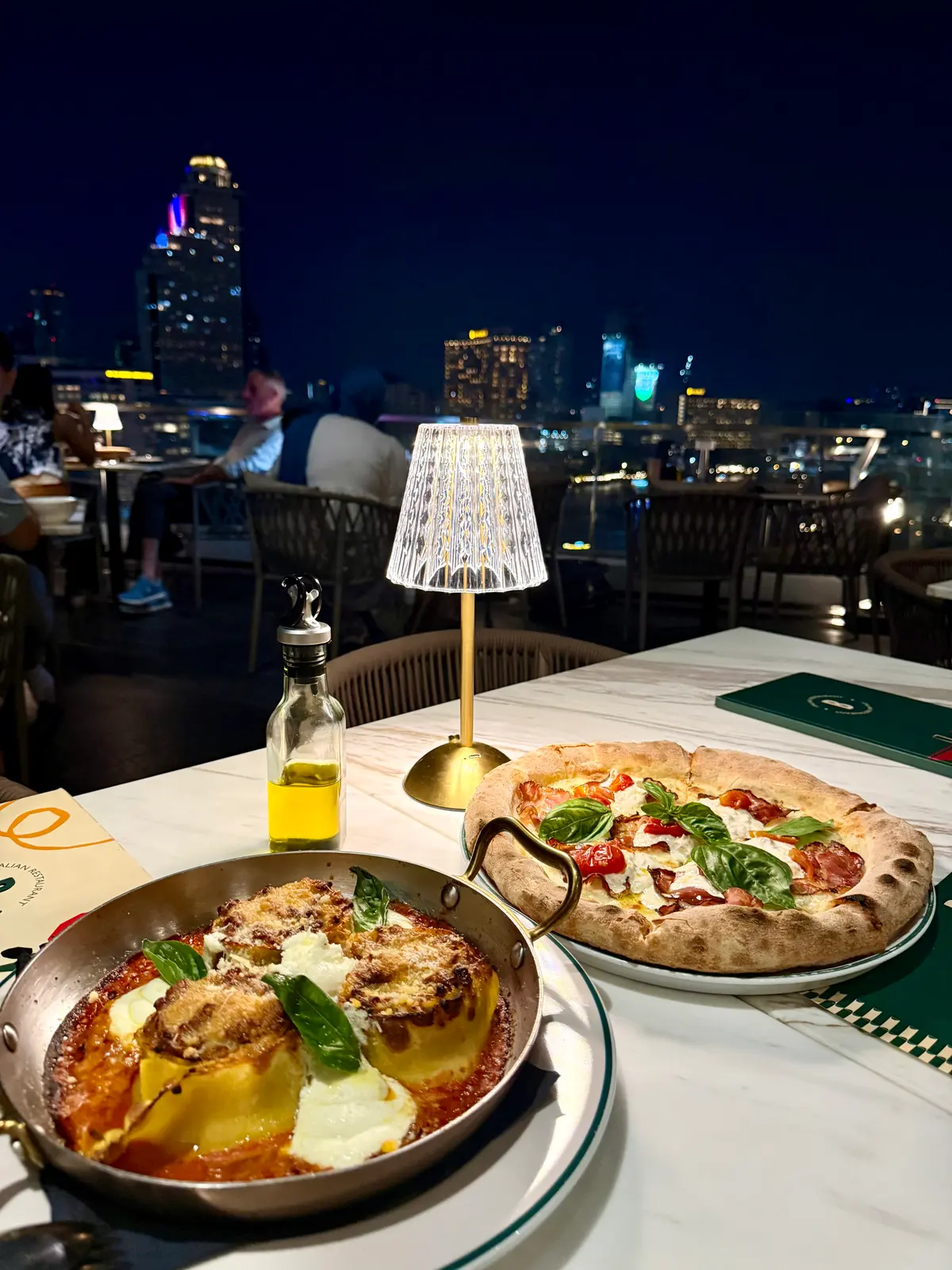 Bianca's lasagna and a wood-fired pizza on a balcony table at night, with State Tower lit up across the river