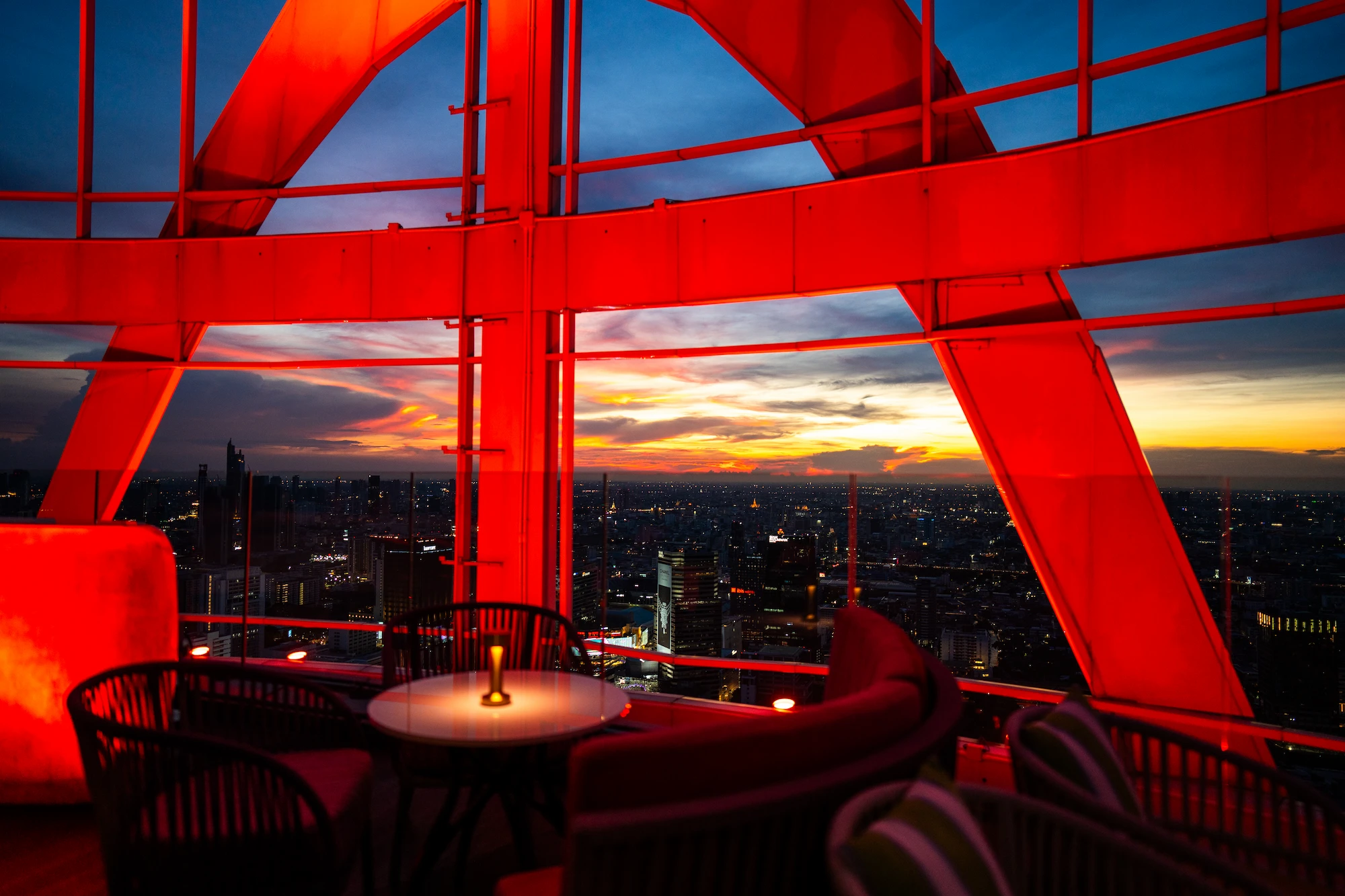 The red-lit steel frame of Cocoa XO's rooftop opening onto a fiery sunset over the Ratchaprasong skyline, Bangkok