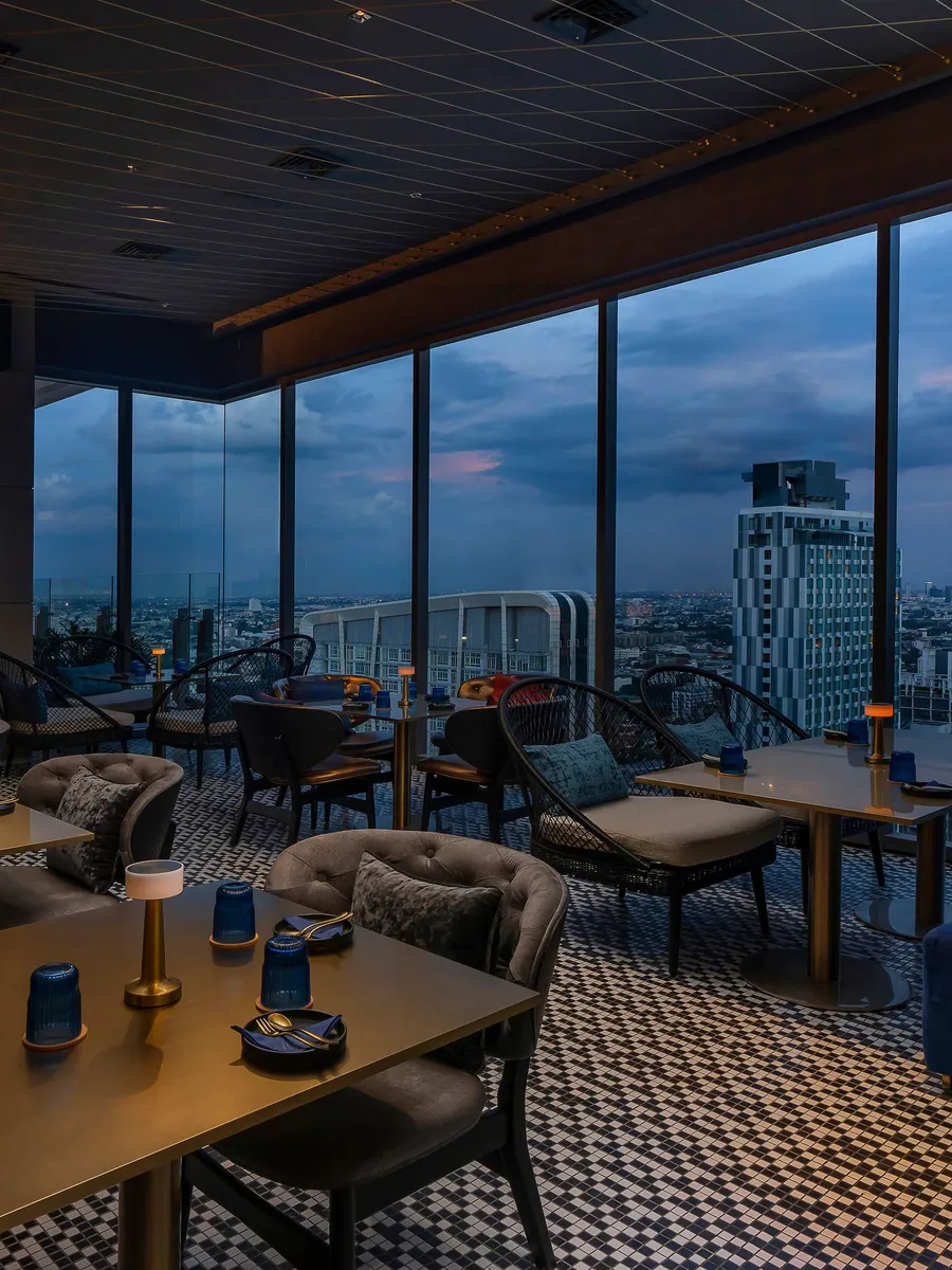 The indoor dining room at LUZ Bangkok Tapas Bar at dusk, blue seating and floor-to-ceiling windows onto the city