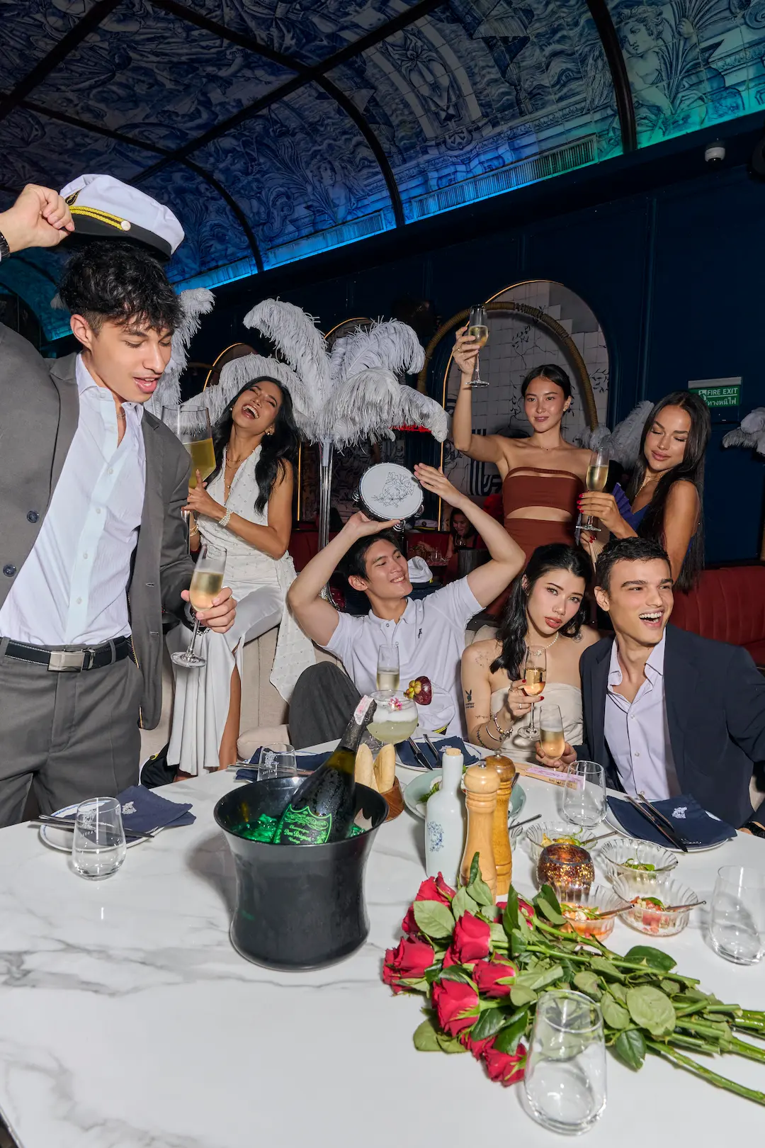 A dressed-up group toasting with champagne around a marble table at Pastel, roses and an ice bucket under the blue-lit Mediterranean ceiling