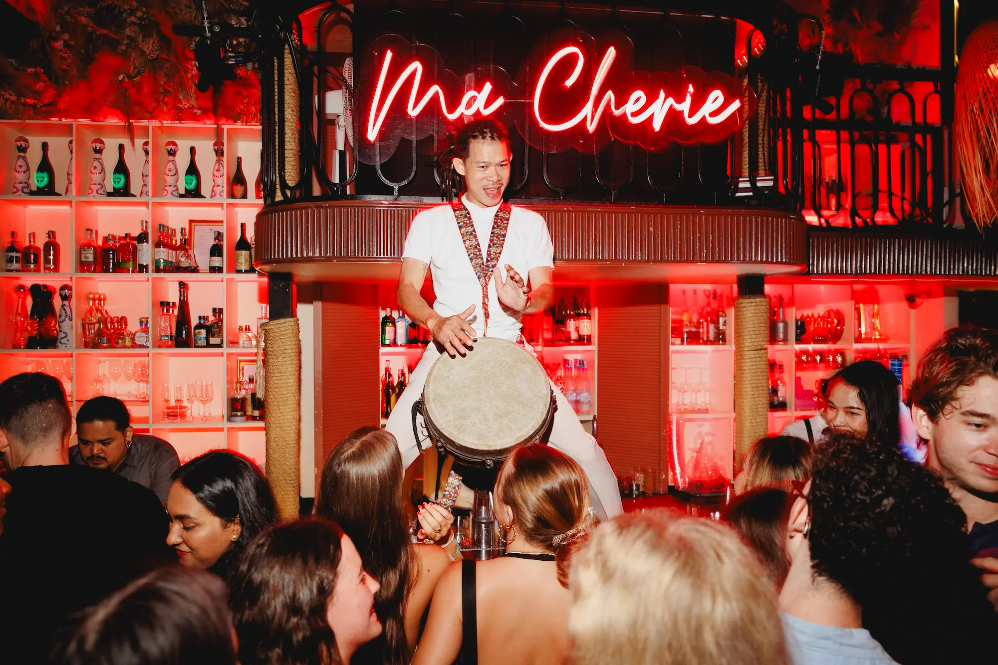 A live percussionist drumming above the crowd under the red neon Ma Cherie sign at Pastel