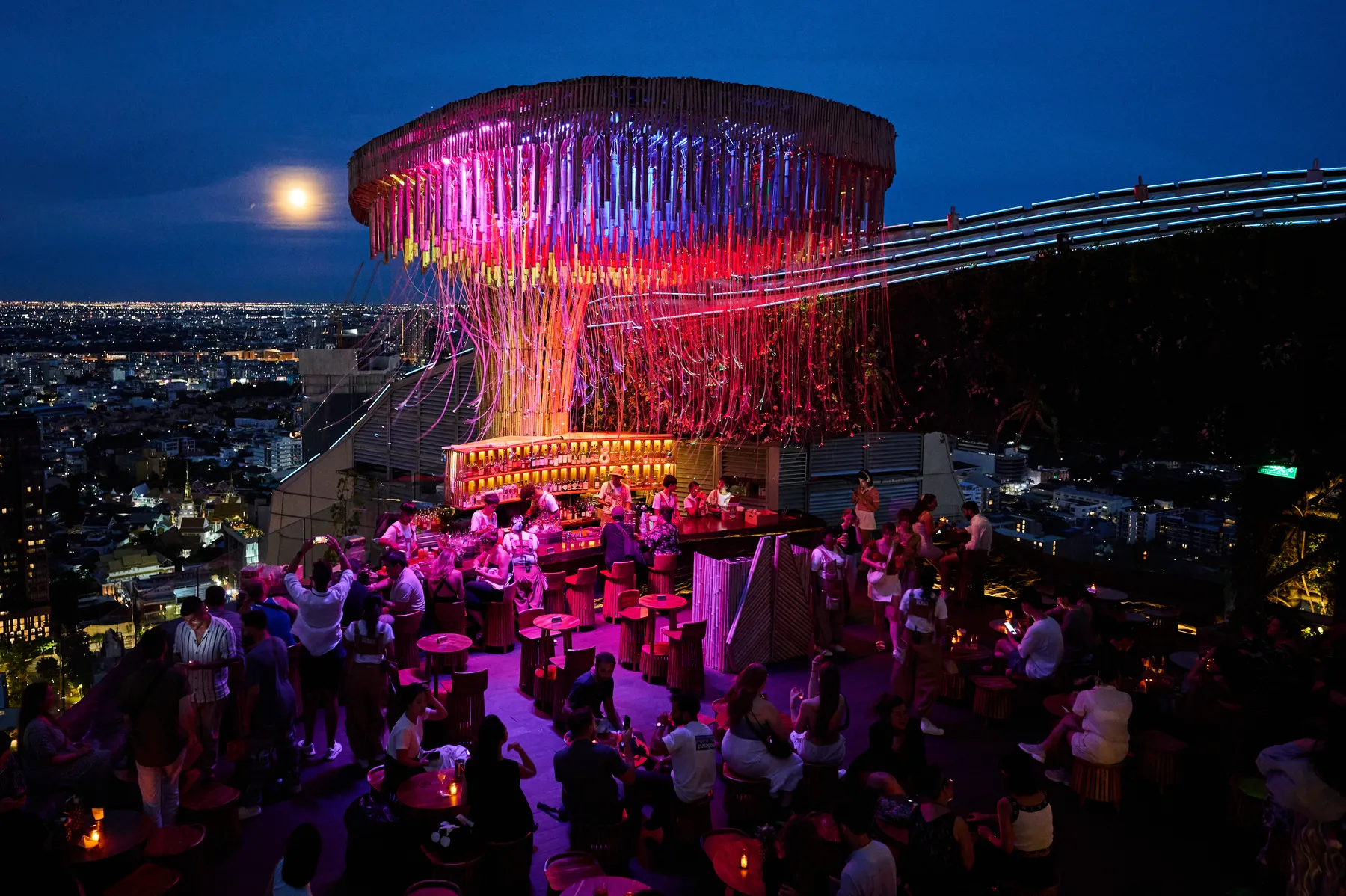 Tichuca's giant canopy glowing pink and red over a busy rooftop terrace at dusk, a full moon rising over the Bangkok skyline