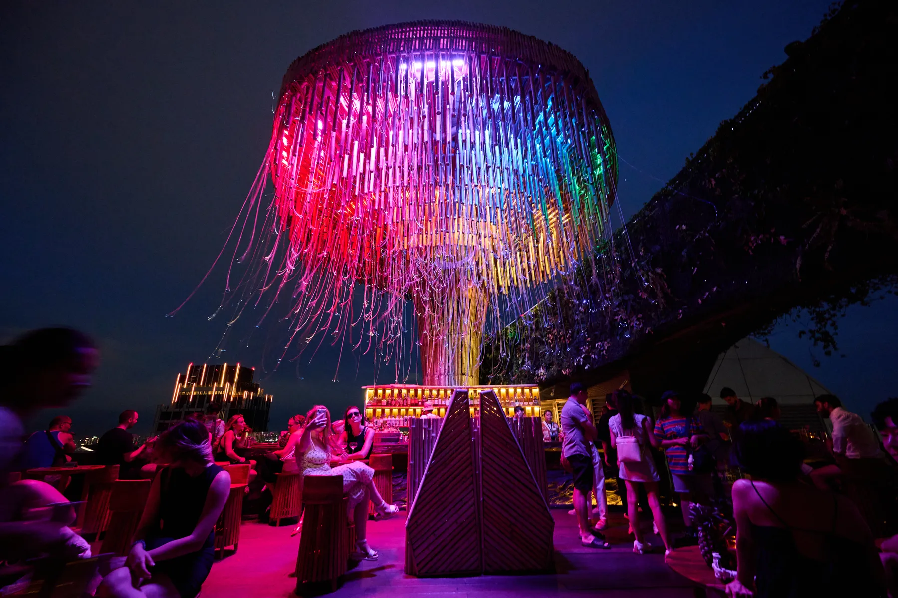 Guests at Tichuca after dark under the canopy lit in pink, blue and green, the city skyline behind