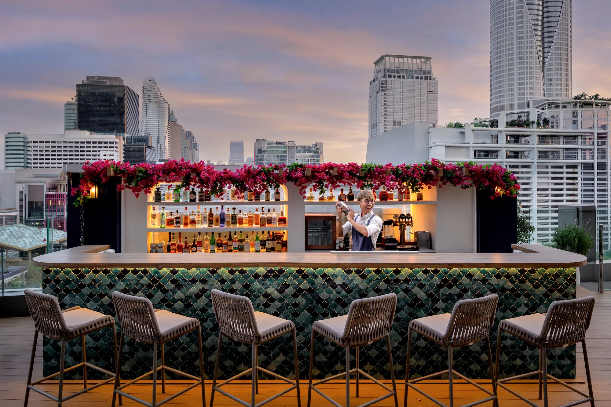 Bartender shaking a cocktail behind View rooftop's bar at dusk, with the Bangkok skyline behind